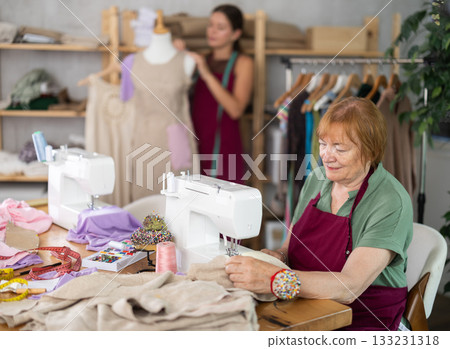 Mature dressmaker sewing clothes against the background of assistant and mannequin Mature dressmaker sewing clothes against the background of assistant and mannequin 133231318