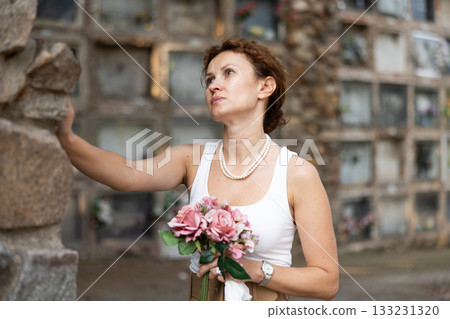 Sad woman in white croptop with bouquet of flowers and handkerchief in hands, stands in cemetery 133231320