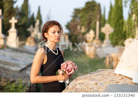 Sad woman in black dress with bouquet of flowers and handkerchief in hands, stands in cemetery 133231340