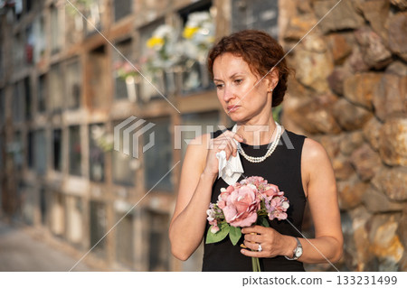 Sad woman in black dress with bouquet of flowers and handkerchief in hands, stands in cemetery 133231499
