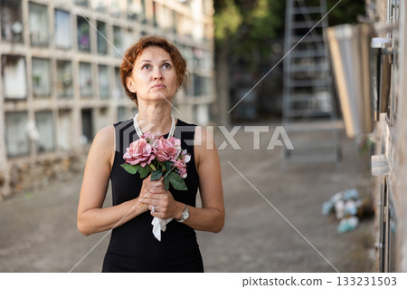 Drieving woman in black dress standing with bouquet by columbarium Drieving woman in black dress standing with bouquet by columbarium 133231503