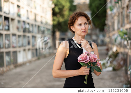 Sad woman in black dress with bouquet of flowers and handkerchief in hands, stands in cemetery 133231606