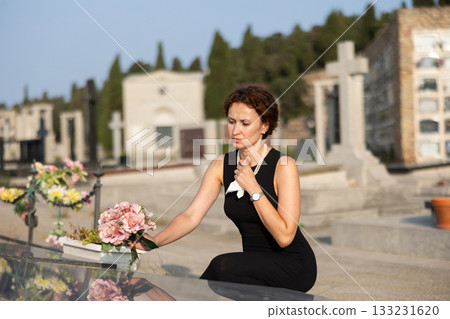 Sorrowful woman in black resting flowers on grave in cemetery 133231620