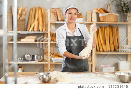 Professional female baker prepares raw yeast dough in bakery, shaping it into baguette 133231646