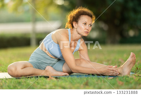 Woman practicing hatha yoga in garden, stretching in an asymmetrical seated asana Parivritta Janu Sirsasana Woman practicing hatha yoga in garden, stretching in an asymmetrical seated asana Parivritta Janu Sirsasana 133231888