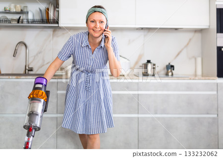 Housewife is cleaning the room - vacuuming and talking on smartphone at same time Housewife is cleaning the room - vacuuming and talking on smartphone at same time 133232062
