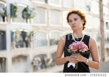 Sorrowful woman in black standing in cemetery with flowers 133232122