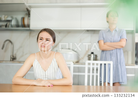Offended young girl sitting at the kitchen table, angry mother standing behind Offended young girl sitting at the kitchen table, angry mother standing behind 133232134