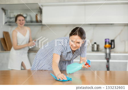 Pleased mother dusting table while daughter washes dishes in kitchen Pleased mother dusting table while daughter washes dishes in kitchen 133232601