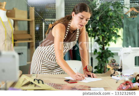 Girl tailor draws pattern on a sheet of paper in sewing workshop 133232605