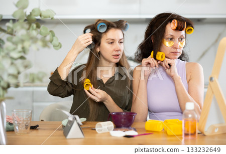 Two adult besties applying hair curlers in front of tabletop mirror 133232649