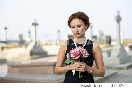 Sad woman in black dress with bouquet of flowers and handkerchief in hands, stands in cemetery 133232653