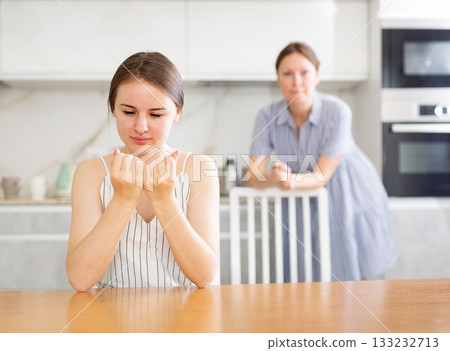Offended young girl sitting at the kitchen table, mother standing behind 133232713