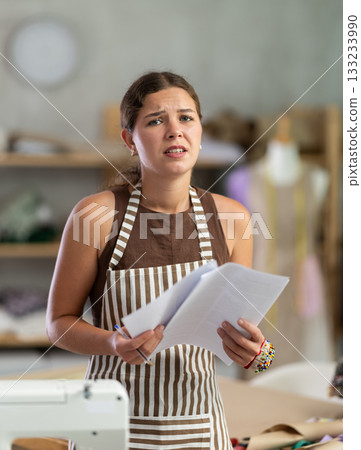 Sad seamstress girl reading financial document in sewing studio interior Sad seamstress girl reading financial document in sewing studio interior 133233990
