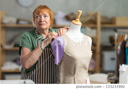 Mature female tailor sews new dress on a mannequin in sewing studio 133233992