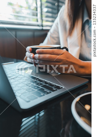 Close-up of a person holding a coffee cup while using a laptop in a contemporary office environment. 133234337