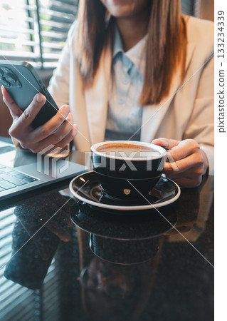 Professional woman in office attire holding a smartphone with a cup of coffee and laptop on a glossy table. Professional woman in office attire holding a smartphone with a cup of coffee and laptop on a glossy table. 133234339