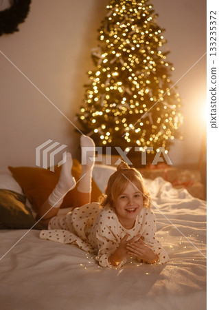 Smiling girl in white dress lying on bed with Christmas tree lights in cozy room 133235372