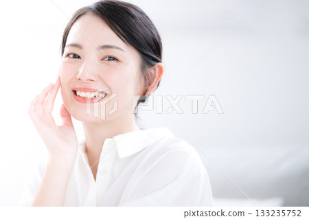 A clean-cut young woman in a white shirt smiles and shows skin care and beauty. Close-up, looking at the camera. 133235752