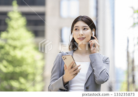A businesswoman in her 30s wearing earphones and holding a smartphone 133235873