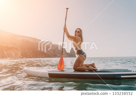 Woman Paddleboarding Ocean Beach - A woman in a bikini kneels on a paddleboard in the ocean. She is holding a paddle and looking at the camera. There are mountains in the background. Woman Paddleboarding Ocean Beach - A woman in a bikini kneels on a paddleboard in the ocean. She is holding a paddle and looking at the camera. There are mountains in the background. 133238552
