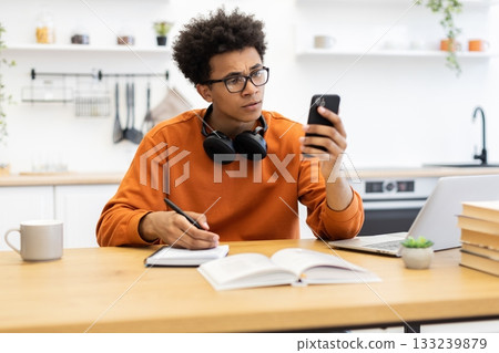 A young man with glasses looks at his phone while working at a table with a laptop, notebook, and coffee cup. 133239879