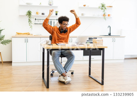 A young man celebrates a victory while working from home, raising his arms in excitement in his kitchen. 133239905