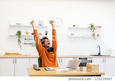 A young man celebrates with arms raised in a bright kitchen setting, possibly after a successful work or study session. 133239907