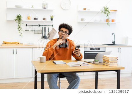 A young man with glasses ponders while holding a phone and pen at a kitchen table with a laptop and books. A young man with glasses ponders while holding a phone and pen at a kitchen table with a laptop and books. 133239917