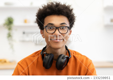 A young man with glasses and headphones around his neck smiles at the camera in a well-lit room. A young man with glasses and headphones around his neck smiles at the camera in a well-lit room. 133239951