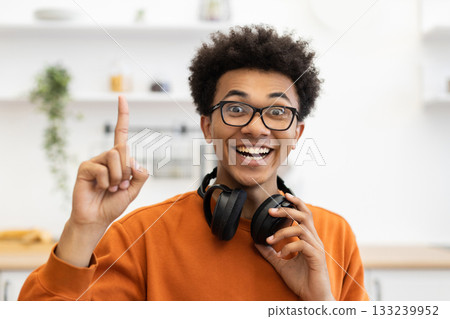 A young Black man with glasses points upwards with a joyful expression, holding headphones. 133239952