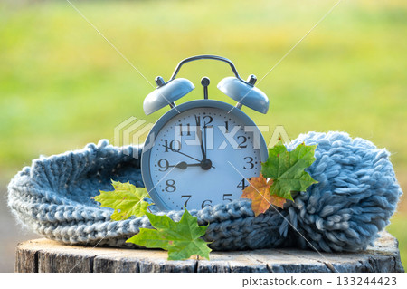 Alarm clock with a knitted hat and autumn leaves on a wooden stump in nature. Alarm clock with a knitted hat and autumn leaves on a wooden stump in nature. 133244423