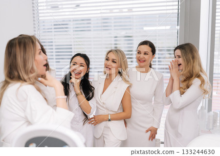 Happy five female healthcare professionals in white uniforms gathered together in modern clinic office, exchanging smiles as they discuss and share their expertise during medical conference. 133244630