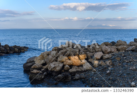 Calm waters embrace rugged stones along the serene coastline at dusk under a gentle sky 133246390