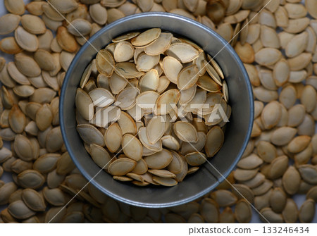 Fresh pumpkin seeds collected in a metal container surrounded by scattered seeds on a wooden surface Fresh pumpkin seeds collected in a metal container surrounded by scattered seeds on a wooden surface 133246434