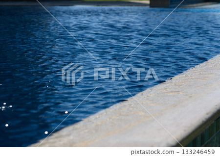 Clear blue water rippling gently against a sunlit pool edge on a warm summer day 133246459