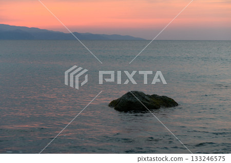 Sunset casting a warm glow over tranquil waters with distant mountains and a lone rock Sunset casting a warm glow over tranquil waters with distant mountains and a lone rock 133246575
