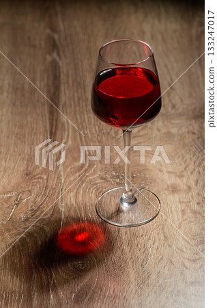 Rich red liquid fills elegant glass casting a playful shadow on warm wooden surface Rich red liquid fills elegant glass casting a playful shadow on warm wooden surface 133247017