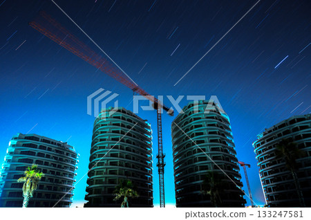 Stars streak across the night sky above newly constructed buildings and cranes in an urban landscape 133247581