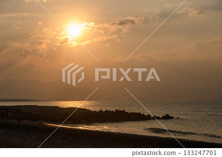 Golden sunset casts warm hues over tranquil shoreline and gentle waves near distant hills 133247701