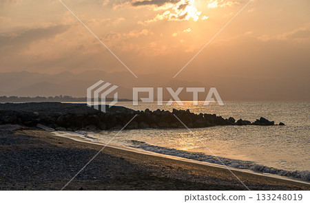 Golden sunset over tranquil beach with distant mountains and gentle waves caressing the shore Golden sunset over tranquil beach with distant mountains and gentle waves caressing the shore 133248019