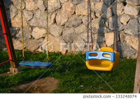 Colorful swings invite joyful play beneath the warm evening sun by a rustic stone wall 133248074
