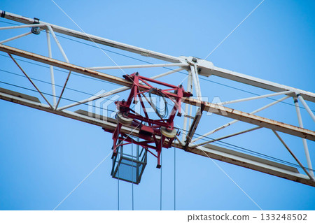 Close-up of White Boom Section with Red Cargo Truck of Tower Crane on Construction Site against Blue Sky in Sunlight. Racks with streaks of Rust, Electricity Wires run horizontally 133248502
