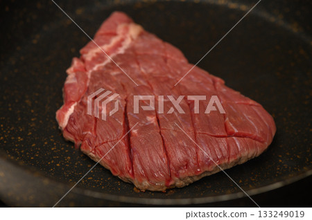 Sizzling preparation of a fresh steak in a black frying pan on a cozy kitchen stovetop 133249019