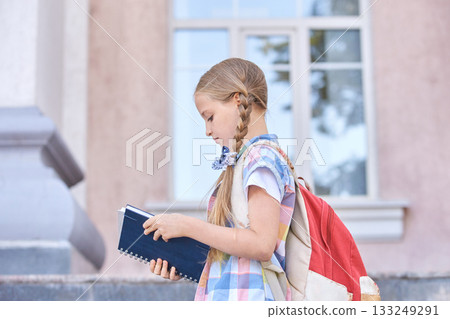 Young caucasian female child with backpack reading notebook outdoors Young caucasian female child with backpack reading notebook outdoors 133249291