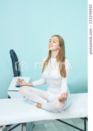 Young caucasian female meditating in white attire on massage table in calm spa 133249485