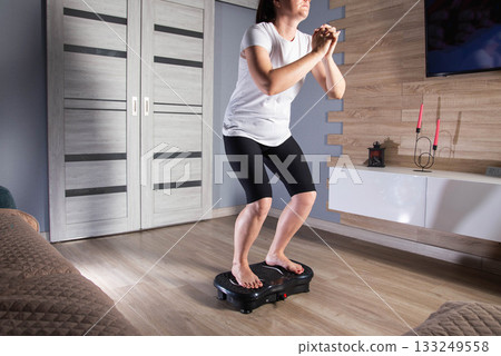 Brunette girl doing sports at home on a modern sports vibration platform. Concept of cellulite elimination and weight loss, background 133249558