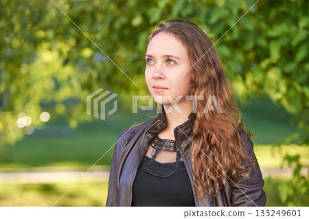 Young caucasian female with long hair in leather jacket standing in sunny park 133249601