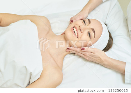Relaxed young caucasian female enjoying facial massage at spa for skin 133250015