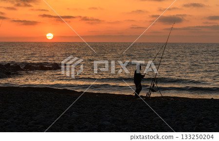 Fisherman casts line at sunset along a tranquil beach with vibrant colors reflecting on the ocean waves Fisherman casts line at sunset along a tranquil beach with vibrant colors reflecting on the ocean waves 133250204